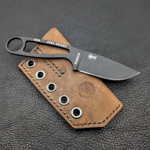 May include: A black ESEE Knives fixed blade knife with a brown leather sheath. The knife has a loop handle and the text "USA" and "ESEE KNIVES" is visible. The sheath has metal grommets and the text "ROWEN" is visible on the knife.