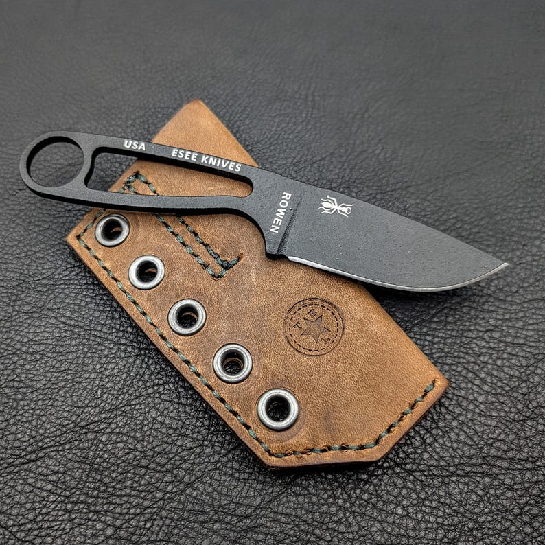 May include: A black ESEE Knives fixed blade knife with a brown leather sheath. The knife has a loop handle and the text "USA" and "ESEE KNIVES" is visible. The sheath has metal grommets and the text "ROWEN" is visible on the knife.