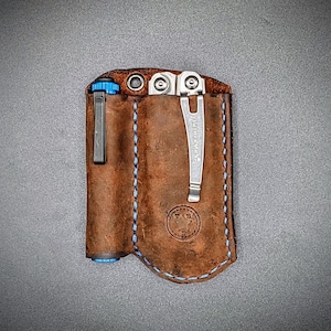May include: A brown leather multi-tool pouch with blue stitching. The pouch has a pocket for a pen or flashlight and a larger pocket for a multi-tool. The multi-tool is a silver Leatherman with a blue handle. The pouch has a star-shaped logo on the front.