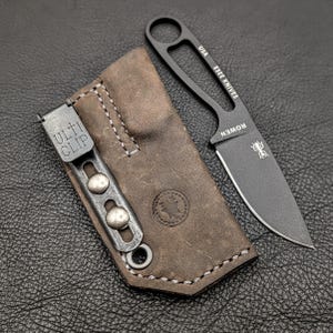 May include: A black ESEE KNIVES fixed blade knife with a brown leather sheath. The knife has a full tang design and a sharp blade. The sheath has a ULTI CLIP and a stitched design.
