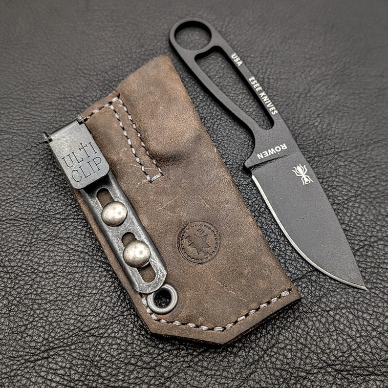 May include: A black ESEE KNIVES fixed blade knife with a brown leather sheath. The knife has a full tang design and a sharp blade. The sheath has a ULTI CLIP and a stitched design.