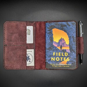 May include: A burgundy leather notebook cover with a pen holder and a pocket for a card. The cover is closed and shows a Field Notes memo book with an image of a rocky archway and a person walking on a beach. The text on the memo book reads "Field Notes Olympic National Park Memo Book 48 Pages / Made in the U.S.A."