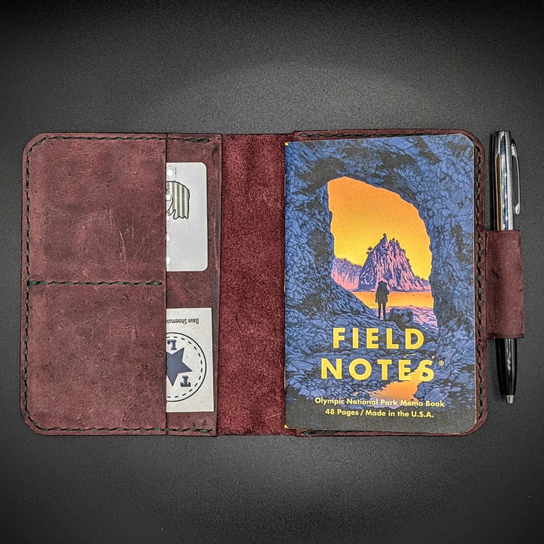 May include: A burgundy leather notebook cover with a pen holder and a pocket for a card. The cover is closed and shows a Field Notes memo book with an image of a rocky archway and a person walking on a beach. The text on the memo book reads "Field Notes Olympic National Park Memo Book 48 Pages / Made in the U.S.A."