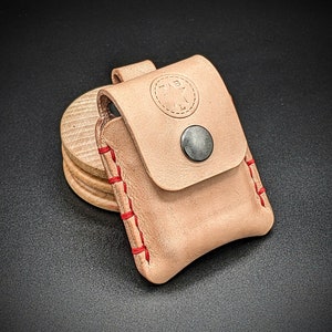 May include: A light brown leather pouch with red stitching. The pouch has a flap with a small metal button closure. The pouch has a star-shaped logo on the front.