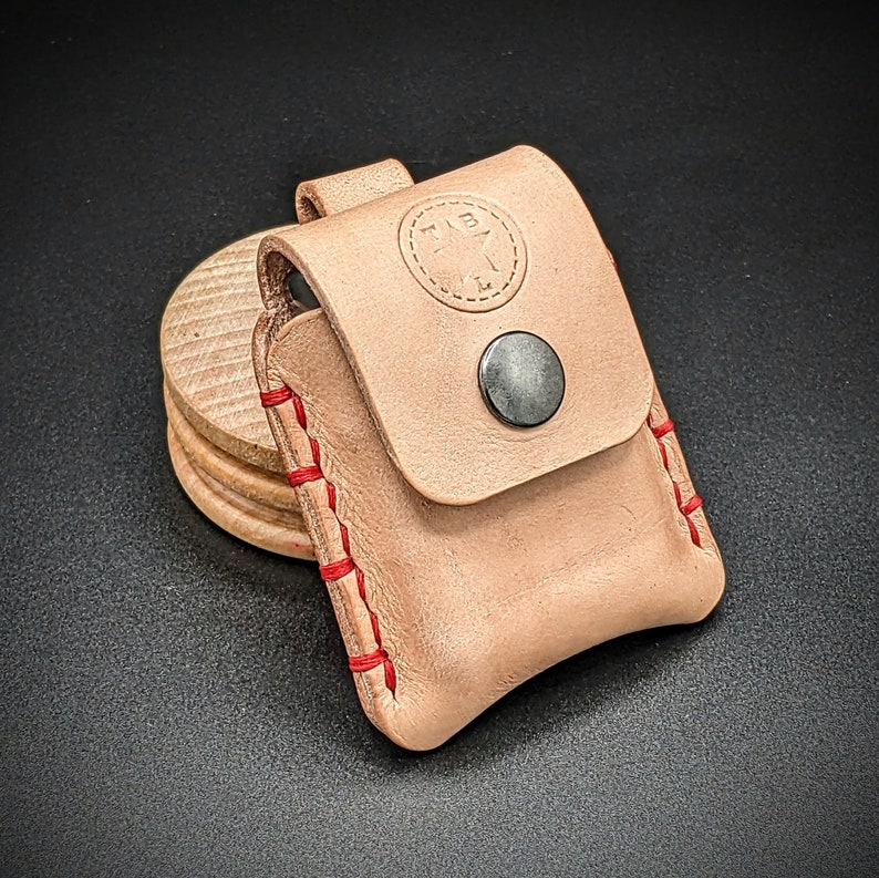 May include: A light brown leather pouch with red stitching. The pouch has a flap with a small metal button closure. The pouch has a star-shaped logo on the front.