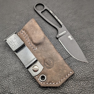 May include: A black ESEE KNIVES fixed blade knife with a brown leather sheath. The knife has a full tang design with a finger hole and the text "USA ESEE KNIVES ROWEN" is visible. The sheath has a metal clip that reads "ULTI CLIP".