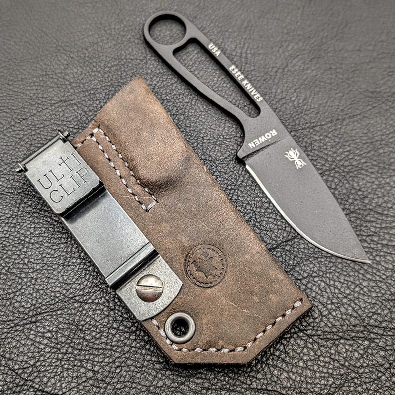 May include: A black ESEE KNIVES fixed blade knife with a brown leather sheath. The knife has a full tang design with a finger hole and the text "USA ESEE KNIVES ROWEN" is visible. The sheath has a metal clip that reads "ULTI CLIP".