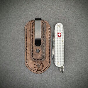 K&ouml;nnte beinhalten: Eine braune Leder-Taschenmesser-Scheide mit einem Metallclip neben einem silbernen Schweizer Taschenmesser. Die Scheide hat ein Sternlogo und die Initialen "TAB". Das Messer hat ein rotes Kreuz-Emblem.