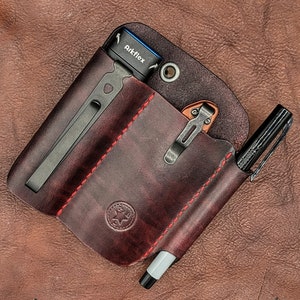 May include: A burgundy leather EDC organizer with red stitching, holding a black pen, a small knife, and a flashlight with "Arkflex" printed on it. The organizer has a metal clip and a stamped logo.