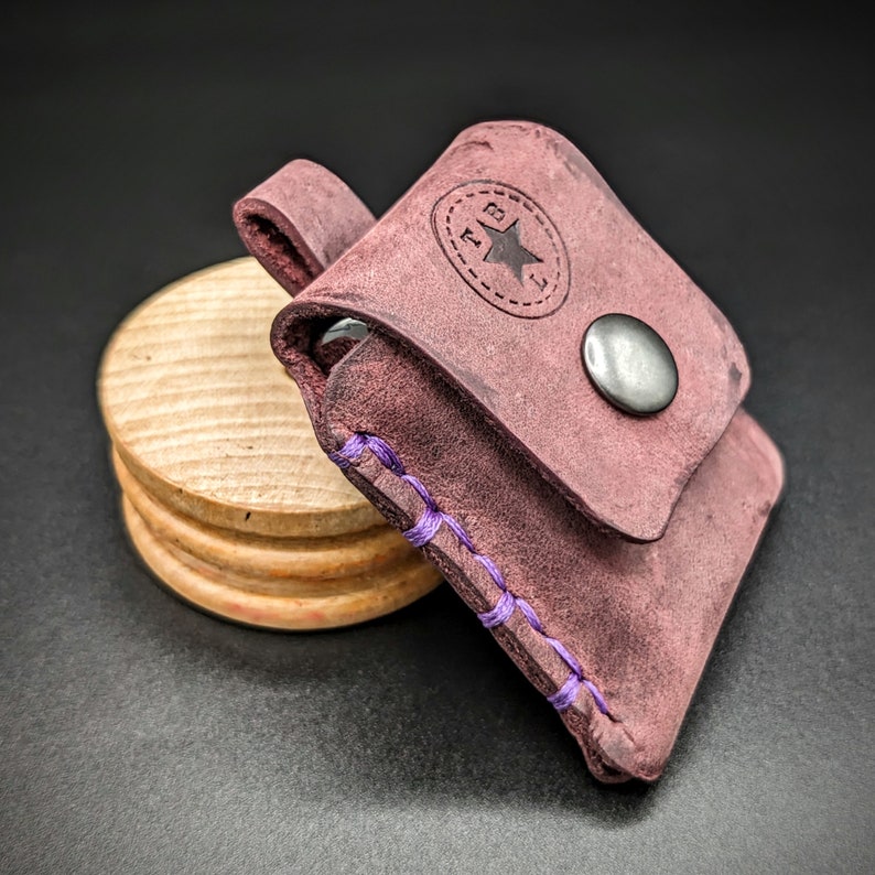 May include: A small, purple leather pouch with a star and the letters "RB" embossed on the front. The pouch has a purple stitching detail and a metal snap closure.