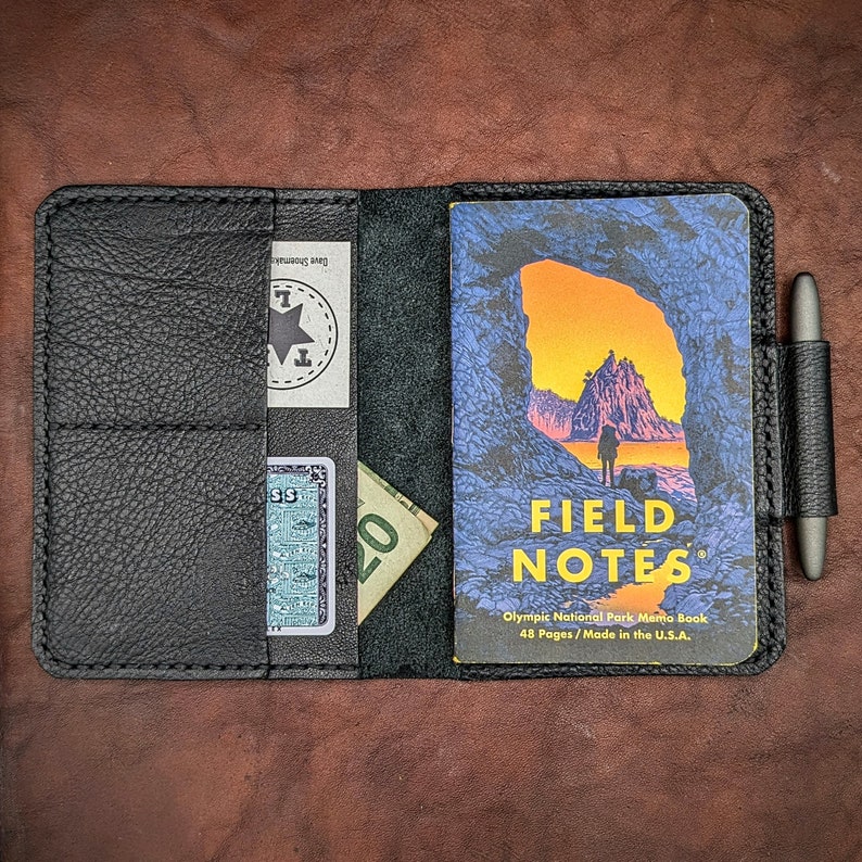 May include: A black leather notebook cover with a pen holder, a pocket for cash and cards, and a Field Notes memo book with a scenic image of a rocky archway and a beach. The memo book cover reads "Olympic National Park Memo Book, 48 Pages, Made in the U.S.A."