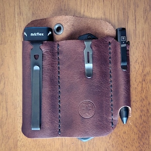 May include: A dark brown leather pocket organizer with three vertical slots. The left slot holds a black Arkflex tool with a metal clip. The center slot contains a black pocket knife with a metal clip. The right slot holds a black pen with a Ti x Knafs clip.