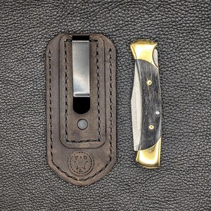 K&ouml;nnte beinhalten: Ein Taschenmesser mit schwarzem Griff und goldenen Akzenten, neben einer braunen Lederscheide mit Metallclip. Das Messer ist geschlossen und zeigt die Klinge und den Griff. Die Scheide hat ein gestempeltes Logo.