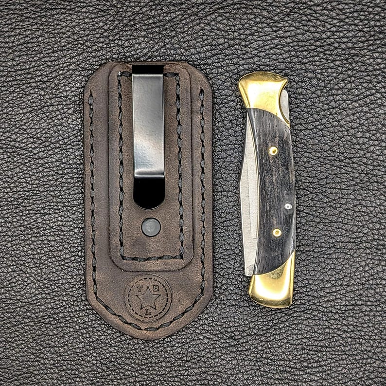 K&ouml;nnte beinhalten: Ein Taschenmesser mit schwarzem Griff und goldenen Akzenten, neben einer braunen Lederscheide mit Metallclip. Das Messer ist geschlossen und zeigt die Klinge und den Griff. Die Scheide hat ein gestempeltes Logo.