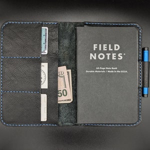 May include: A black leather notebook cover with blue stitching. The cover has multiple pockets and slots for cards and cash. The cover is open to reveal a black Field Notes notebook with the text "FIELD NOTES" and "64-Page Note Book Durable Materials / Made in the U.S.A."