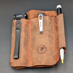 May include: A brown leather pocket organizer with red stitching. It features a black clip, a silver clip with the word "Kershaw", and a pen. The organizer has the text "Arkflex" and a logo with the letters "TAB".