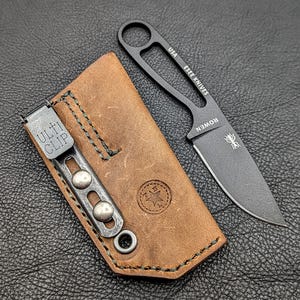 May include: A black ESEE KNIVES fixed blade knife with a brown leather sheath. The knife has a curved blade and a handle with a finger hole. The sheath has a metal clip that reads "ULTI CLIP". The knife and sheath are on a dark surface.