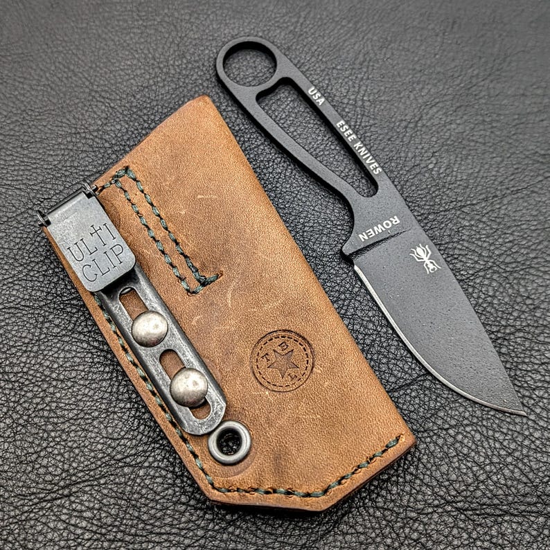 May include: A black ESEE KNIVES fixed blade knife with a brown leather sheath. The knife has a curved blade and a handle with a finger hole. The sheath has a metal clip that reads "ULTI CLIP". The knife and sheath are on a dark surface.