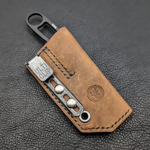 May include: A small, black knife with a ring handle is partially inserted into a brown leather sheath. The sheath has a metal clip with the text "ULTI CLIP" and is stitched with green thread. The sheath also has a stamped logo.