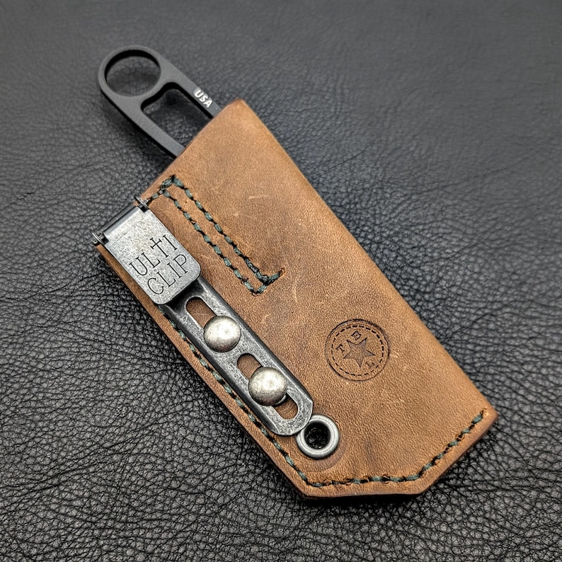 May include: A small, black knife with a ring handle is partially inserted into a brown leather sheath. The sheath has a metal clip with the text "ULTI CLIP" and is stitched with green thread. The sheath also has a stamped logo.
