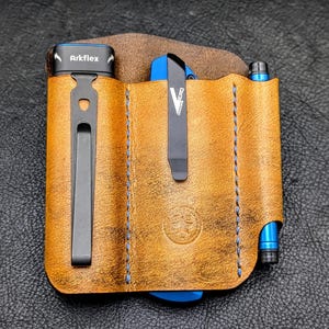 May include: A tan leather pocket organizer with three vertical slots. The left slot holds a black Arkflex tool with a metal clip. The center slot contains a blue folding knife with a black clip. The right slot holds a blue pen with a metal tip.