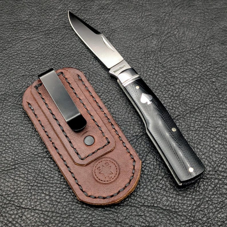 K&ouml;nnte beinhalten: Ein Klappmesser mit schwarzem Griff und silberner Klinge, neben einer braunen Lederscheide mit schwarzem Clip. Der Messergriff hat ein wei&szlig;es Pik-Symbol.