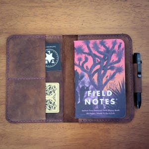 May include: A brown leather notebook cover with purple stitching, holding a Field Notes Joshua Tree National Park memo book with a pink and purple desert graphic, two small cards, and a black pen secured in a side loop on the right side.