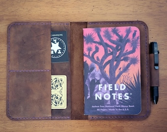 May include: A brown leather notebook cover with purple stitching, holding a Field Notes Joshua Tree National Park memo book with a pink and purple desert graphic, two small cards, and a black pen secured in a side loop on the right side.