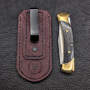 K&ouml;nnte beinhalten: Ein Taschenmesser mit schwarzem Griff und goldenen Akzenten, neben einer weinroten Lederscheide mit schwarzem Clip. Das Messer hat eine silberne Klinge und ist teilweise ge&ouml;ffnet. Die Scheide hat ein gepr&auml;gtes Logo.