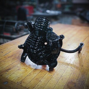 May include: Black cast iron hand crank food grinder with a decorative design. The grinder has a handle and a large wheel with a hole in the center.