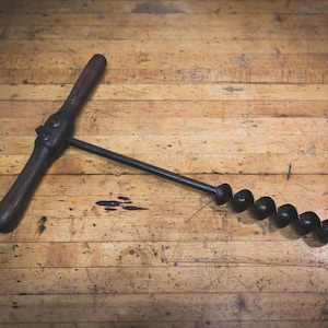 May include: A vintage wooden-handled drill bit with a black metal spiral bit. The drill bit is lying on a wooden surface.
