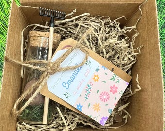 May include: A small gift box contains a glass bottle with a cork stopper, a miniature rake, and a tag tied with twine. The tag has the name "Emanuel" and the date "12 ottobre 2023" with floral illustrations.