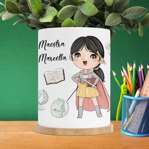 Può includere: Tazza in ceramica bianca con un'illustrazione di un cartone animato di un'insegnante che indossa un mantello da supereroe e tiene un libro. Il testo "Maestra Marcella" è stampato sulla tazza.