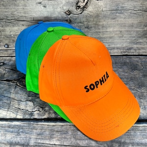 Può includere: Tre cappellini da baseball impilati uno sopra l'altro. Il cappellino superiore è arancione con il nome "Sophia" ricamato in nero. Il cappellino intermedio è verde e il cappellino inferiore è blu.