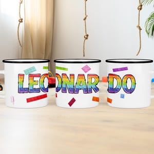 Può includere: Tre tazze di smalto bianco con bordi neri. Ogni tazza ha un nome scritto in arcobaleno. I nomi sono "LEO", "NAR" e "DO".