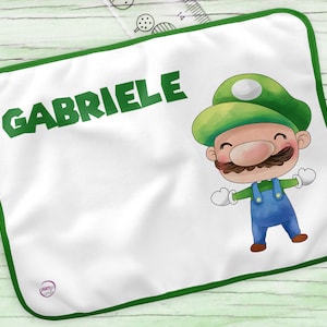 Può includere: Coperta personalizzata bianca con bordo verde. La coperta presenta il nome "Gabriele" in lettere verdi e un personaggio dei cartoni animati con un cappello verde e una salopette blu.