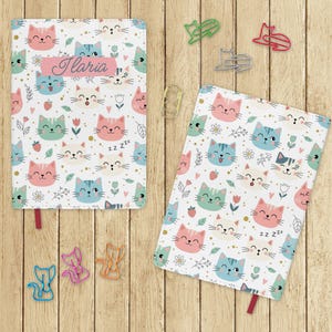 Può includere: Due quaderni con sfondo bianco ricoperti da un motivo di facce di gatti colorate, fiori e fragole. Un quaderno ha il nome "Ilaria" su uno striscione rosa. Fermagli a forma di gatto sono su una superficie di legno.