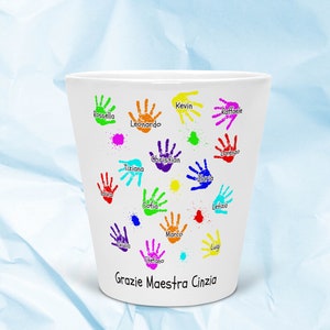 Può includere: Tazza in ceramica bianca con impronte di mani colorate e nomi di bambini, tra cui "Grazie Maestra Cinzia" in fondo.