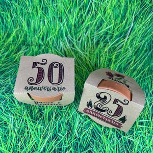 Può includere: Due scatole di cartone marrone con un piccolo vaso di terracotta all'interno di ciascuna. Le scatole sono decorate con i numeri 50 e 25, la parola "aniversario" e un motivo floreale.