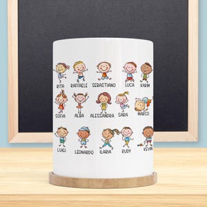 Può includere: Una tazza in ceramica bianca con illustrazioni di cartoni animati di bambini con nomi come Rita, Raffaele e Luca. La tazza ha una base in legno e si trova su uno sfondo di lavagna.