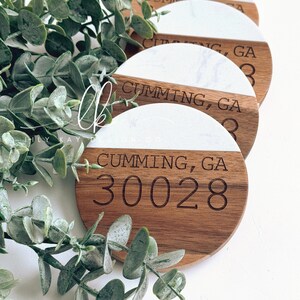 May include: Set of four round wooden coasters with a white marble top. The coasters are engraved with the text "Cumming, GA 30028".