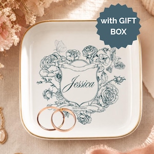 May include: A square white ceramic trinket dish with a gold-toned rim, featuring a dark teal floral illustration, a central shield design with the name Jessica, the date 3/27/1990, and a dark teal star-shaped badge with the text with GIFT BOX.