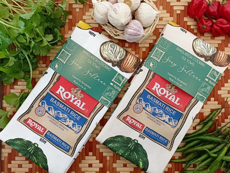 May include: Two white tea towels with a design featuring a bag of Royal Basmati Rice, text, and a green leaf. The towels are labeled "Bangladesh Cooking Tea Towel" and "Sony Sultana". Fresh vegetables surround the towels.