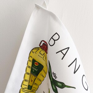 May include: White kitchen towel with colorful illustrations of food items and the word "BANG" in black. The towel features a yellow vegetable oil bottle, green chili peppers, and other food graphics.