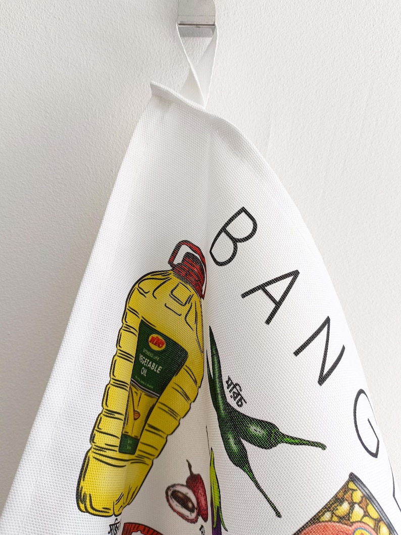 May include: White kitchen towel with colorful illustrations of food items and the word "BANG" in black. The towel features a yellow vegetable oil bottle, green chili peppers, and other food graphics.
