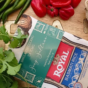 May include: A tea towel with the text "Bangladesh Cooking Tea Towel Sony Sultana" and "Royal Basmati Rice" branding. The towel is surrounded by fresh ingredients like green chilies, red bell peppers, cilantro, and garlic.