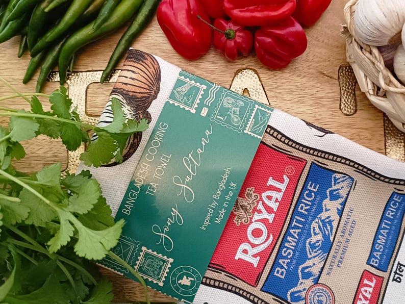 May include: A tea towel with the text "Bangladesh Cooking Tea Towel Sony Sultana" and "Royal Basmati Rice" branding. The towel is surrounded by fresh ingredients like green chilies, red bell peppers, cilantro, and garlic.