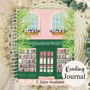 May include: A spiral-bound reading journal with a colorful illustration of a bookstore facade. The cover features a pink brick building with green trim, arched windows, and the text "The Book Shop". The journal is labeled "Reading Journal" and states "2 Sizes Available".