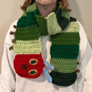 May include: A green, red, and blue crocheted scarf shaped like a caterpillar. The caterpillar has a red head with two black eyes and a black mouth.