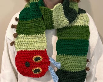 May include: A green, red, and blue crocheted scarf shaped like a caterpillar. The caterpillar has a red head with two black eyes and a black mouth.